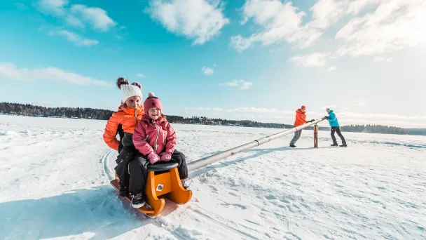 Kaksi lasta talvivaatteissa jäällä napakelkassa järven jäällä aikuisten työntäessä.