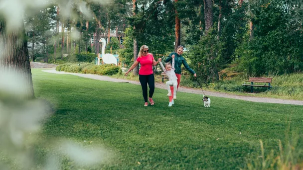 Kaksi ihmistä kävelee nurmikolla koiran kanssa, puisto taustalla.