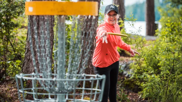 Hymyilevä tyttö heittää kiekkoa frisbeegolf-koriin