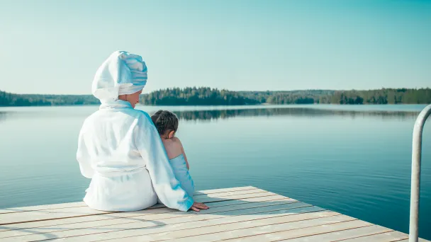 Nainen valkoisessa kylpytakissa ja tyttö pyyhe ympärillään istuvat laiturilla katsellen tyyntä, kesäistä järveä.