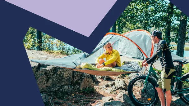 Järven rannalla puihin on kiinnitetty tentsile, jossa nainen istuu ja hymyilee fatbikea pitelevälle miehelle.