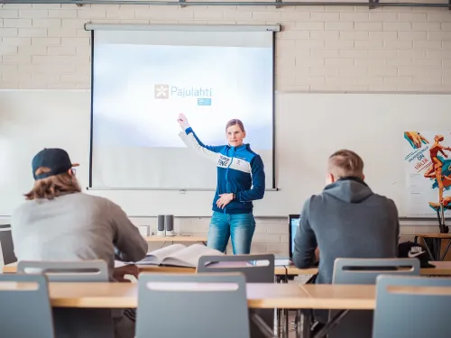 Luokkahuone, opettaja näyttää diaa, opiskelijat istuvat pöydissä.