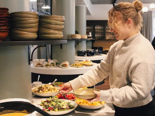 Nainen ottaa salaattia seisovasta pöydästä ravintolassa, hymyillen.