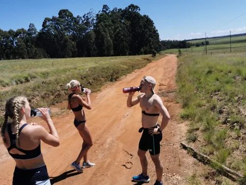 Kolme ihmistä juoksevat hiekkatiellä, juoden vettä auringonpaisteessa.