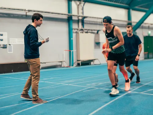 Athletes running on an indoor track while a coach times them.