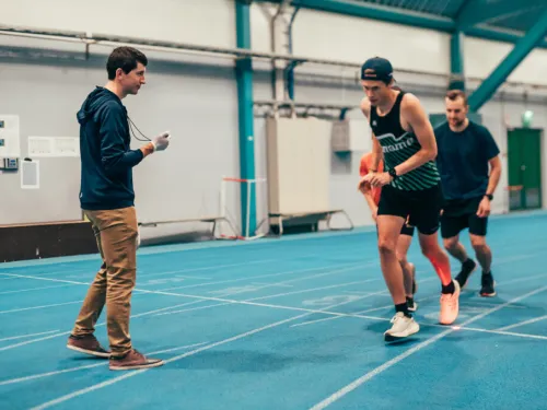 Athletes running on an indoor track while a coach times them.