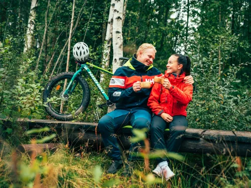 Pari istuu metsässä pyörän kanssa, hymyilee toisilleen.