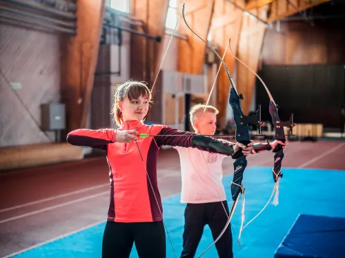 Child and teen practicing archery indoors on a blue mat.