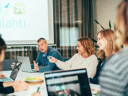 Iloinen ryhmä kokouksessa neuvotteluhuoneessa, kannettavat tietokoneet pöydällä.