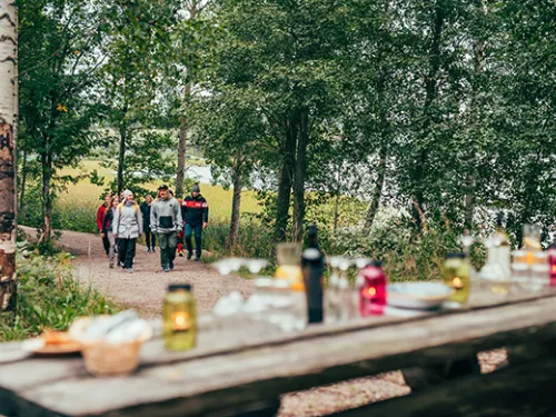 Picnic table with jars in a forest; people walking on trail in background.