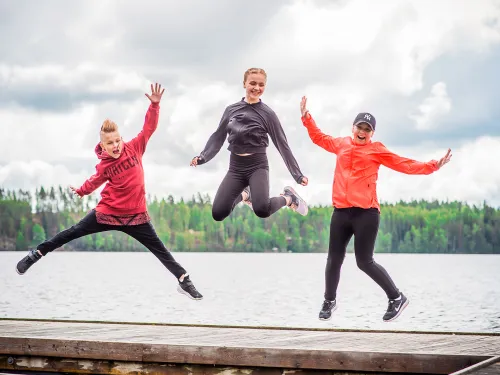 Kolme ihmistä hyppää laiturilla järven edessä, taustalla metsää.