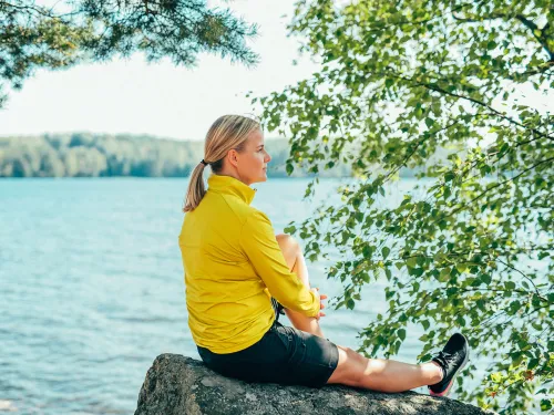 Nainen istuu kalliolla järven rannalla, taustalla puita.