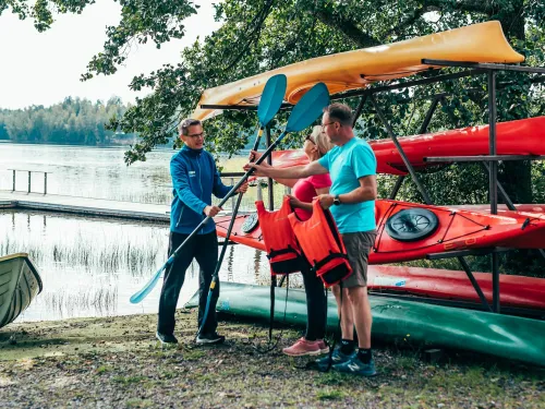 Three people by kayaks at a lakeside, holding paddles and life jackets.