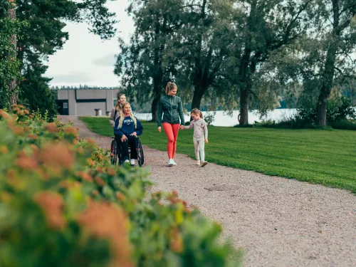 Kolme ihmistä kävelyllä puistossa järven rannalla, yksi henkilö pyörätuolissa.