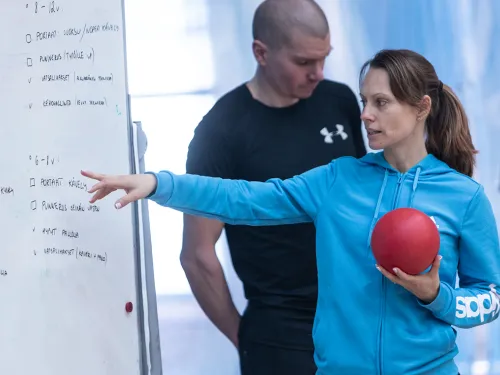 Naishenkilö sinisessä hupparissa osoittaa taululle, pallo kädessä.