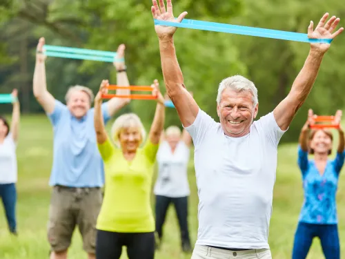 Ryhmä seniori-ikäisiä jumppaa nurmikolla jumppakuminauhoilla ja hymyilee.