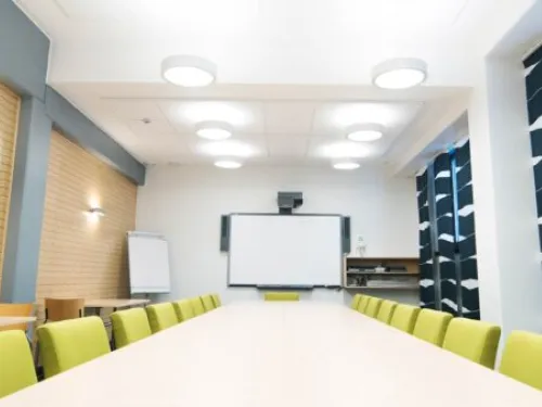 Kokoushuone, pitkät pöydät ja keltaiset tuolit, valkoinen taulu edessä.