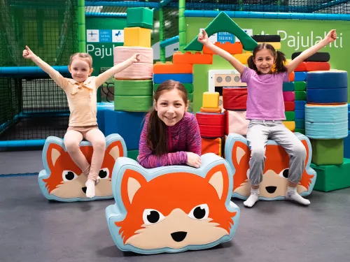 Kolme lasta leikkipaikalla, hymyillen ja istuen kettuaiheisten tyynyjen päällä.