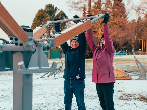 Senioripariskunta kuntoilee Pajulahden rannassa ulkokuntoilulaitteilla hymyillen.