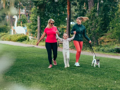 Pajulahden rantanurmikolla kävelee leikkisästi mummo, tyttö ja nainen, koira kävelee heidän vierellään.