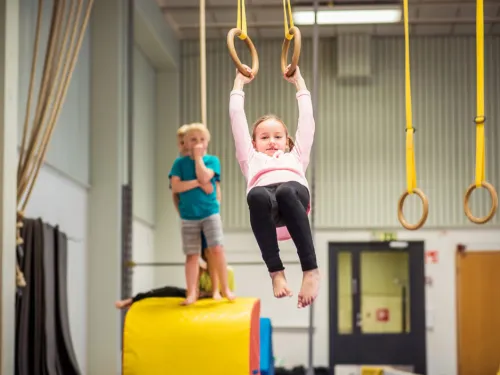 Lapsi roikkuu voimistelurenkaissa, toinen seisoo keltaisella matolla.
