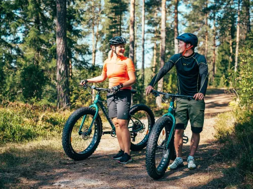 Kaksi henkilöä pyöräilemässä metsäpolulla, aurinkoinen päivä.