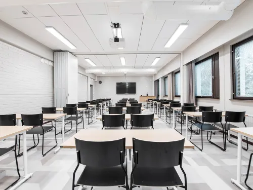 Tyhjä luokkahuone, rivissä mustia tuoleja ja pöytiä, seinillä valkoisia tiiliä ja ikkunoita.