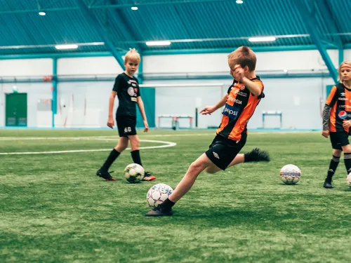 Kolme lasta pelaa jalkapalloa sisätiloissa sinisellä kentällä.