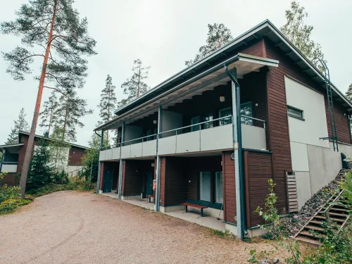 Kaksikerroksinen puutalo metsän keskellä, ympärillä hiekkaa ja puita.