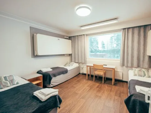 Kaksi vuoteellinen huone, jossa pöytä ja suuri ikkuna harmailla verhoilla.