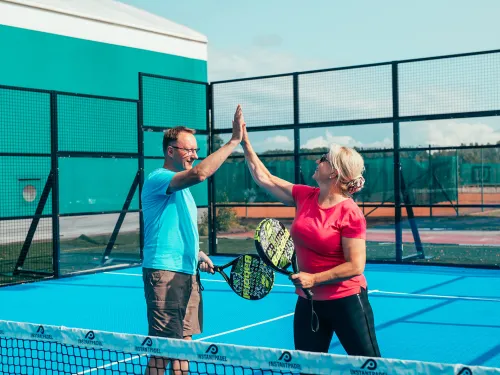 Kaksi henkilöä pelaa padelia ja antaa ylävitosta kentällä.