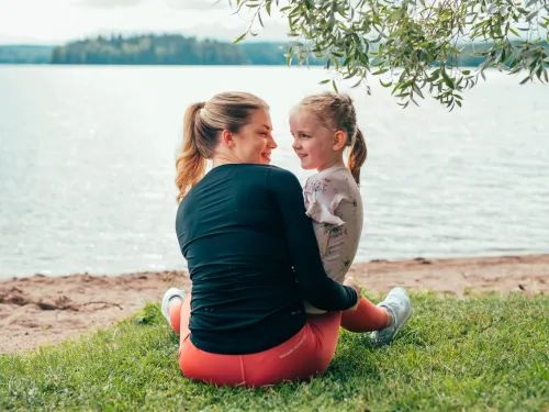 Äiti ja lapsi istuvat nurmikolla järven rannalla kesäisenä päivänä.