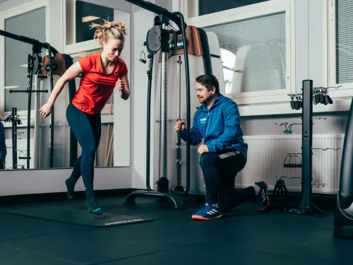 Nainen treenaa salilla, valmentaja kannustaa vieressä.