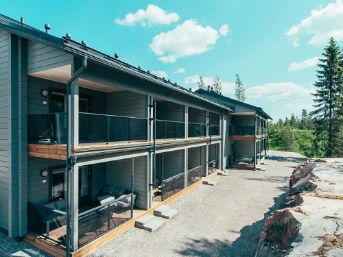 Kaksikerroksinen harmaa puutalo metsämaisemassa, aurinkoinen sää.