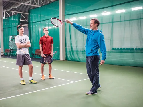 Tennisharjoitus hallissa, valmentaja ohjaa kahta nuorta pelaajaa.