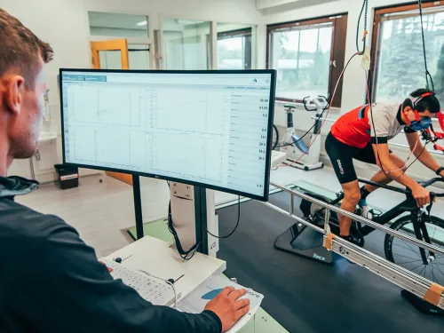 Sisätiloissa pyöräilevä henkilö testissä, taustalla näyttö ja analysoiva tutkija.