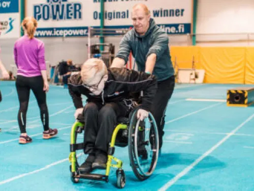 Mies työntää naista pyörätuolissa urheiluhallissa. Taustalla ihmisiä treenaamassa.