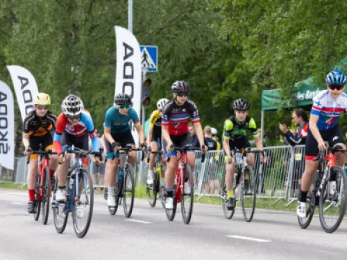 Kilpailijat polkevat pyörillään maantiellä, taustalla puita ja kylttejä.