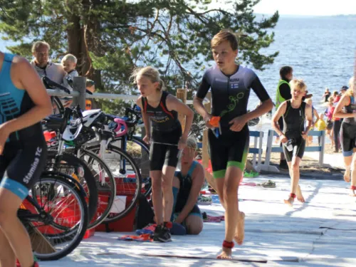 Lasten triathlonkilpailu järven rannalla, pyöriä näkyy taustalla.