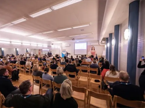 Valokuva konferenssista, jossa on paljon istuvia ihmisiä ja esiintyjä lavalla.