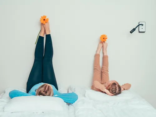 Kaksi henkilöä makaa sängyllä, jalat nostettuina seinää vasten, oranssi kukka jaloissaan.