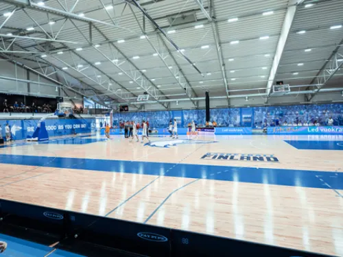 Koripallokenttä sisähallissa, katsojia taustalla.