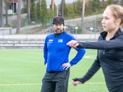 Coach in a blue jacket observing a woman in black running on a field.