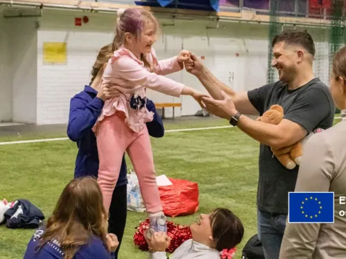 Aikuiset auttavat hymyilevää lasta seisomaan ulkonaurheilutilassa.