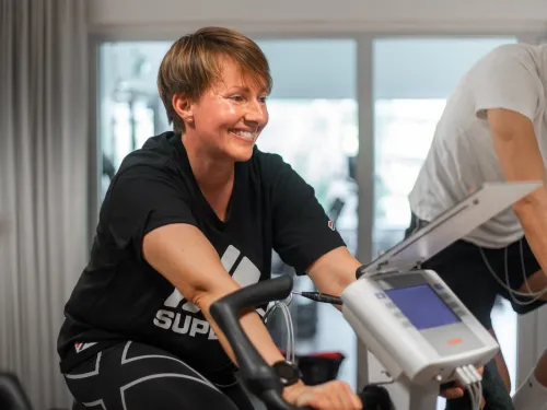 Hymyilevä nainen kuntopyörällä valoisassa huoneessa, vierellään toinen pyöräilijä.