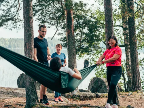 Neljä ihmistä puiden keskellä järven rannalla, yksi makaa riippumatossa.