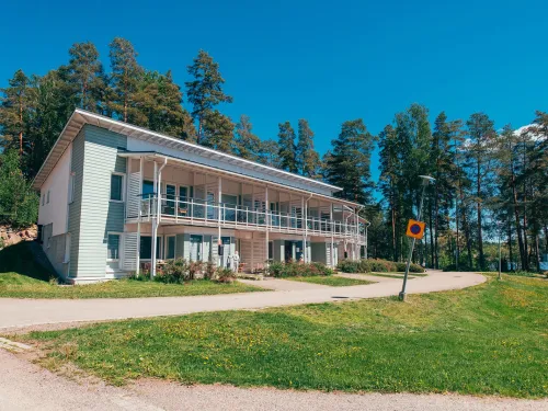 Kaksikerroksinen talo mäntymetsän edessä aurinkoisella päivällä.