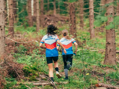 Kaksi henkilöä juoksee metsässä kilpa-asuissa.