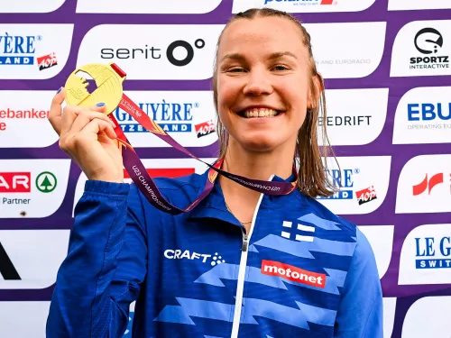 Tähtiurheilija sinisessä asussa, hymyilee ja pitää kultaista mitalia taustallaan sponsoreiden logoja.