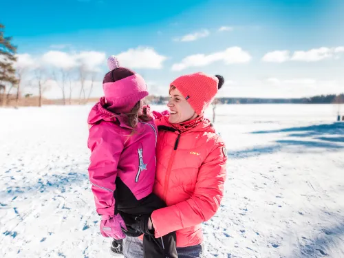 Hymyilevä nainen pitelee sylissään lasta, taustalla kaunis, talvinen järvimaisema.